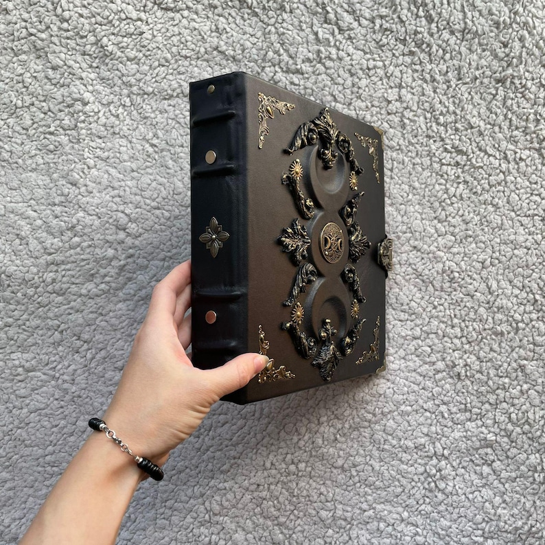 K&ouml;nnte beinhalten: Ein braunes und schwarzes ledergebundenes Buch mit aufwendigen goldfarbenen Metallverzierungen. Das Buch hat einen dekorativen Verschluss und Eckakzente. Der Buchr&uuml;cken ist schwarz mit einem goldfarbenen Blumendesign. Das Buch wird von einer Person gehalten.