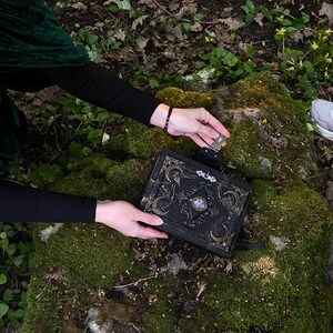May include: Black, ornate book with gold accents and a purple gemstone center, held over a mossy rock. The book has intricate designs and a clasp. The background features forest foliage and a person's hands.