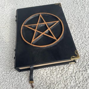 May include: Black leather-bound journal with a gold pentagram embossed on the front cover. The journal has a gold clasp and a black ribbon bookmark.