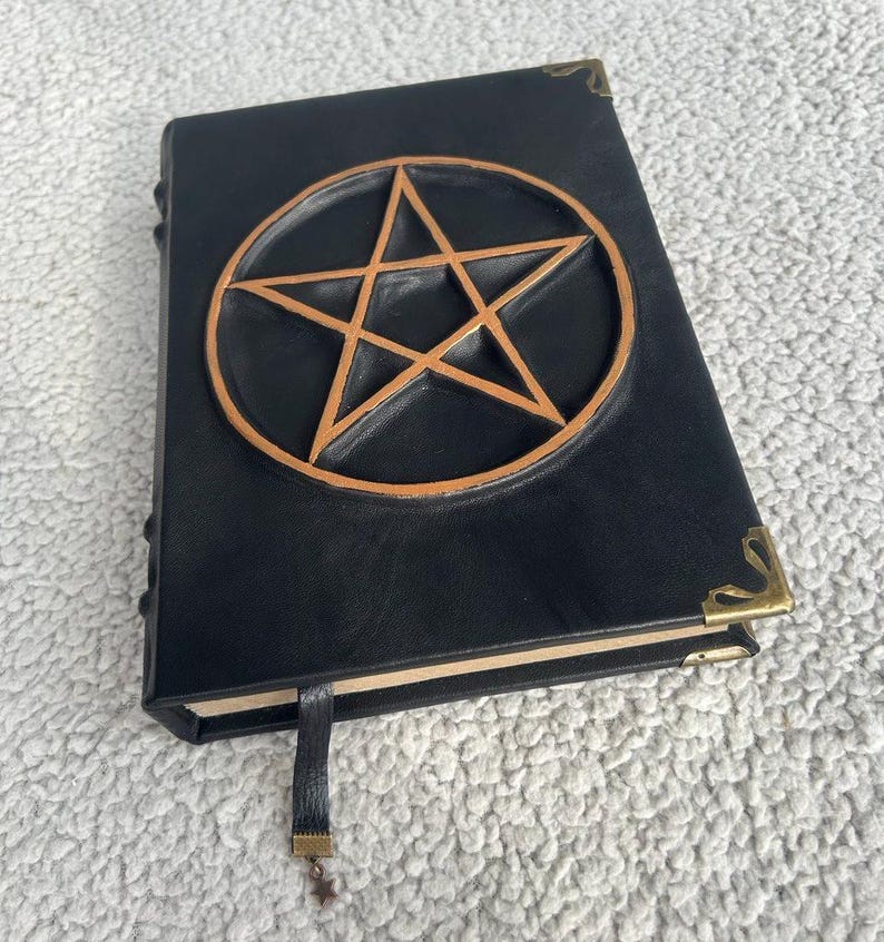May include: Black leather-bound journal with a gold pentagram embossed on the front cover. The journal has a gold clasp and a black ribbon bookmark.