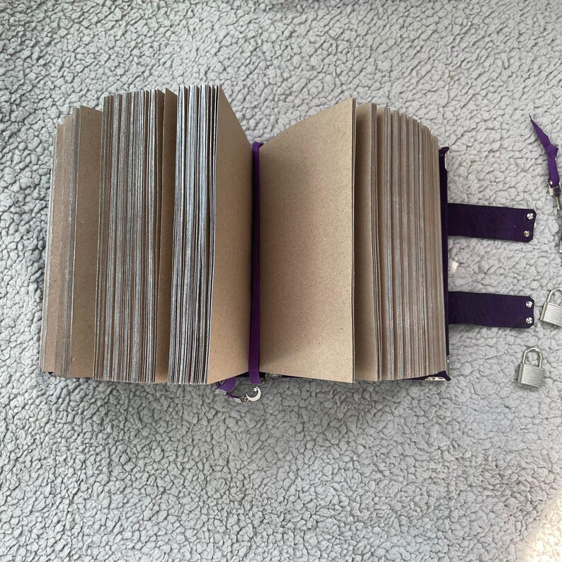 May include: An open journal with kraft paper pages, bound in dark purple suede with matching straps and silver locks. A purple ribbon bookmark is visible. The journal is on a grey, textured surface.