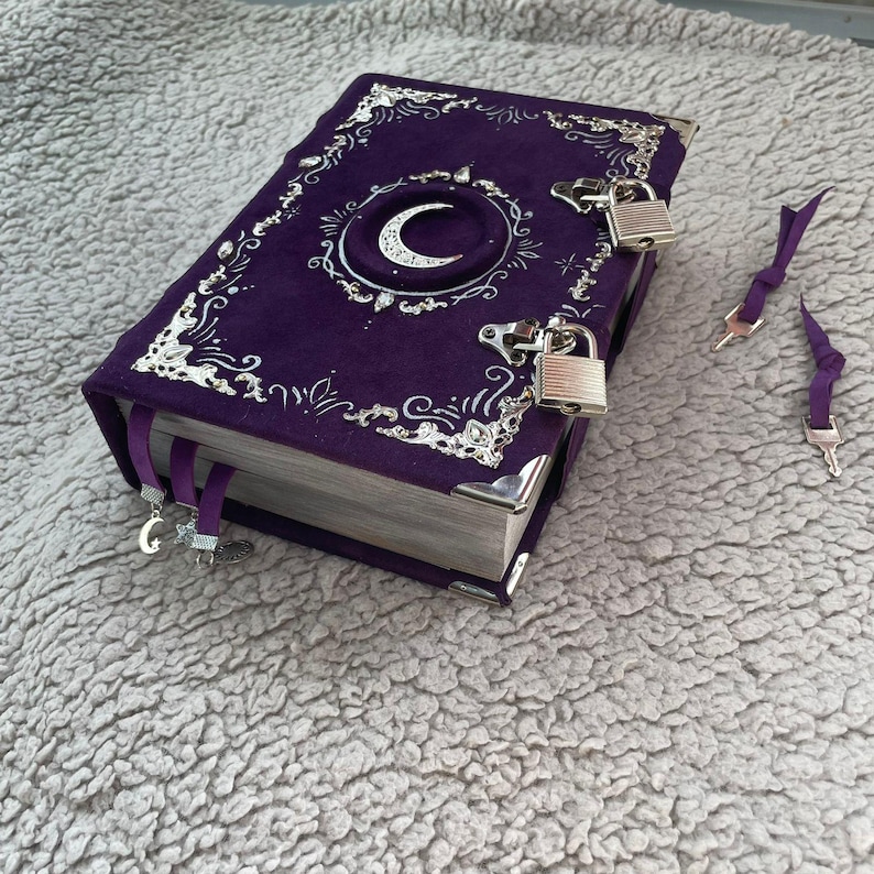 May include: A deep purple journal with silver detailing and a crescent moon motif. The cover is adorned with silver filigree and corner protectors. Two silver padlocks secure the journal, with two keys. Purple ribbon bookmarks are included.