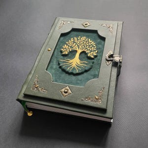 Puede incluir: Un diario de cuero verde con un diseño de árbol dorado en relieve en la portada. El diario presenta esquinas decorativas de metal y un cierre de broche. Se ve una cinta marcapáginas verde.