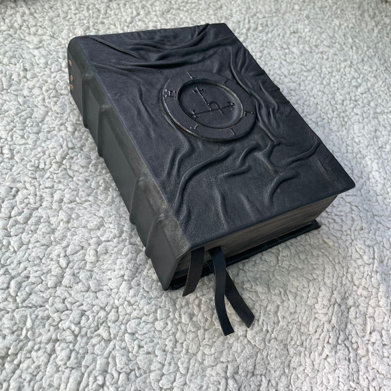 May include: A black leather-bound book with a textured cover and a circular embossed design. The book has gold-colored rivets along the spine and black ribbon bookmarks. The cover features a clock-like emblem.