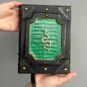 Pode incluir: Um di&aacute;rio encadernado em couro preto com uma capa de couro de crocodilo verde em relevo e um design de cobra dourada. O di&aacute;rio tem um marcador de fita preta com um amuleto de lagarto dourado.