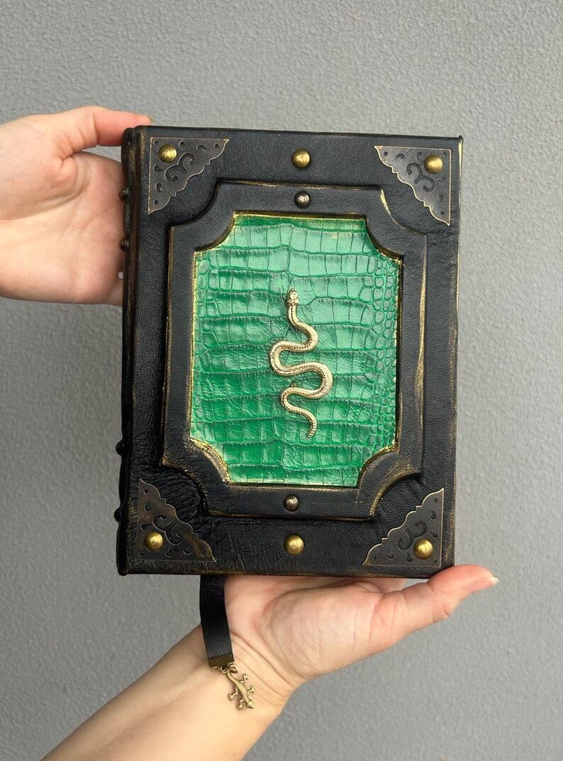 Pode incluir: Um di&aacute;rio encadernado em couro preto com uma capa de couro de crocodilo verde em relevo e um design de cobra dourada. O di&aacute;rio tem um marcador de fita preta com um amuleto de lagarto dourado.