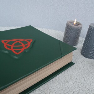 May include: A green leather-bound book with a red Celtic knot symbol on the cover. The book is closed and sitting on a white fluffy surface. Two gray candles are in the background.