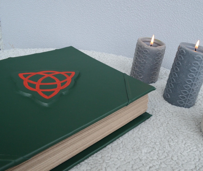 May include: A green leather-bound book with a red Celtic knot symbol on the cover. The book is closed and sitting on a white fluffy surface. Two gray candles are in the background.