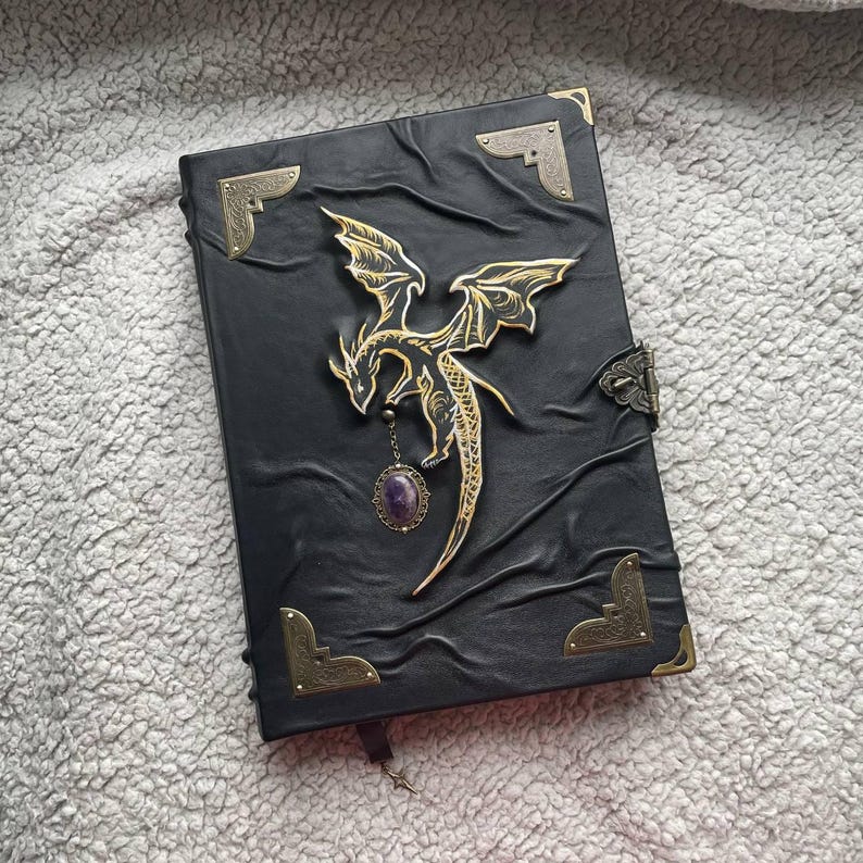 Pu&ograve; includere: Diario in pelle nera con un'applicazione a forma di drago dorato e un ciondolo di ametista. Il diario ha angoli protettivi in ottone e una chiusura. Il drago &egrave; dettagliato con ali spiegate. Un taccuino a tema fantasy.