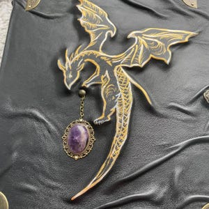 Pu&ograve; includere: Diario in pelle nera con un disegno di drago dorato e bianco. Un ciondolo con pietra preziosa viola pende dall'artiglio del drago. Il diario ha angoli decorativi in metallo.