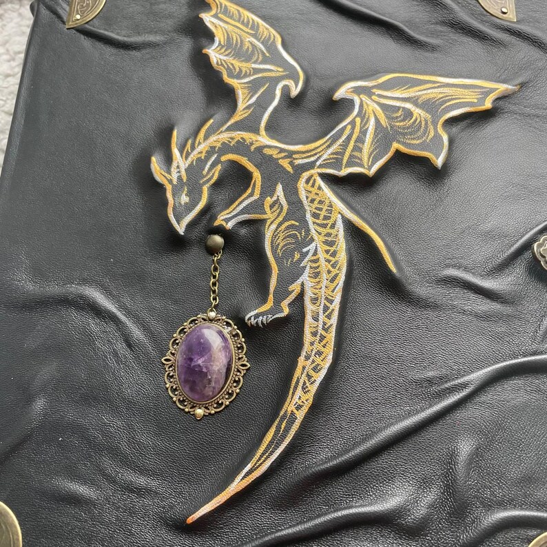 Pu&ograve; includere: Diario in pelle nera con un disegno di drago dorato e bianco. Un ciondolo con pietra preziosa viola pende dall'artiglio del drago. Il diario ha angoli decorativi in metallo.