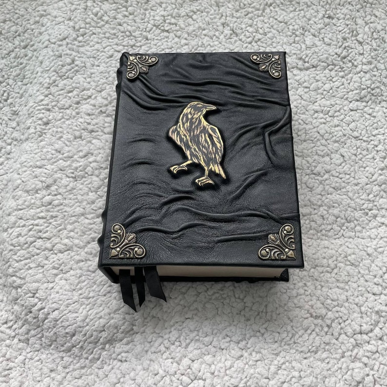 Pu&ograve; includere: Un diario in pelle nera con un emblema di corvo dorato e dettagli decorativi agli angoli. Il diario ha una copertina strutturata e un segnalibro a nastro nero. Un taccuino unico ed elegante per scrivere o disegnare.