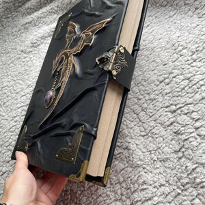 Pu&ograve; includere: Diario rilegato in pelle nera con un design a drago. La copertina presenta un drago dorato in rilievo, un ciondolo con pietra viola e protezioni angolari in metallo ornate. Una chiusura decorativa assicura il diario.