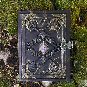 May include: A black, decorative journal with ornate gold and bronze accents. The cover features a central oval with a purple stone, surrounded by intricate designs and crescent moon details. A metal clasp secures the journal.