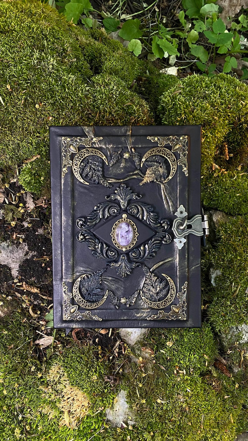 May include: A black, decorative journal with ornate gold and bronze accents. The cover features a central oval with a purple stone, surrounded by intricate designs and crescent moon details. A metal clasp secures the journal.