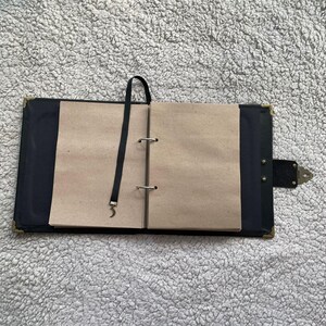 K&ouml;nnte beinhalten: Offenes schwarzes Notizbuch mit leeren, hellbraunen Seiten. Das Notizbuch hat ein schwarzes Leseband und einen Verschluss. Die Ecken sind mit goldfarbenem Metall verst&auml;rkt. Das Notizbuch ist ge&ouml;ffnet und zeigt die Innenseiten.