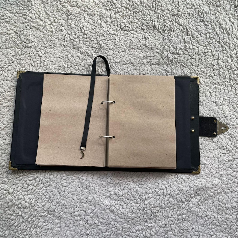 K&ouml;nnte beinhalten: Offenes schwarzes Notizbuch mit leeren, hellbraunen Seiten. Das Notizbuch hat ein schwarzes Leseband und einen Verschluss. Die Ecken sind mit goldfarbenem Metall verst&auml;rkt. Das Notizbuch ist ge&ouml;ffnet und zeigt die Innenseiten.