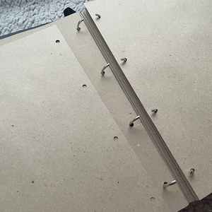 May include: An open black binder with several pages of light brown paper. The pages are held together by metal rings. The binder is on a light-colored, textured surface.