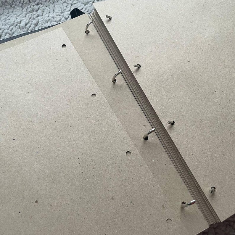 May include: An open black binder with several pages of light brown paper. The pages are held together by metal rings. The binder is on a light-colored, textured surface.