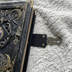 May include: A black leather journal with ornate brass corner protectors and embellishments. The cover features a purple gemstone and a crescent moon design. A black leather strap with a brass clasp secures the journal.