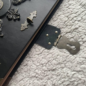 May include: Black leather journal with ornate metal clasp and decorative accents. The journal features a keyhole clasp and brass-colored hardware. The cover has embossed floral and scroll designs, creating a vintage aesthetic.