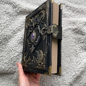 May include: A black leather-bound journal with ornate gold-colored metal accents and a purple gemstone. The cover features embossed designs and a clasp closure. The journal's corners are reinforced with gold-colored metal.