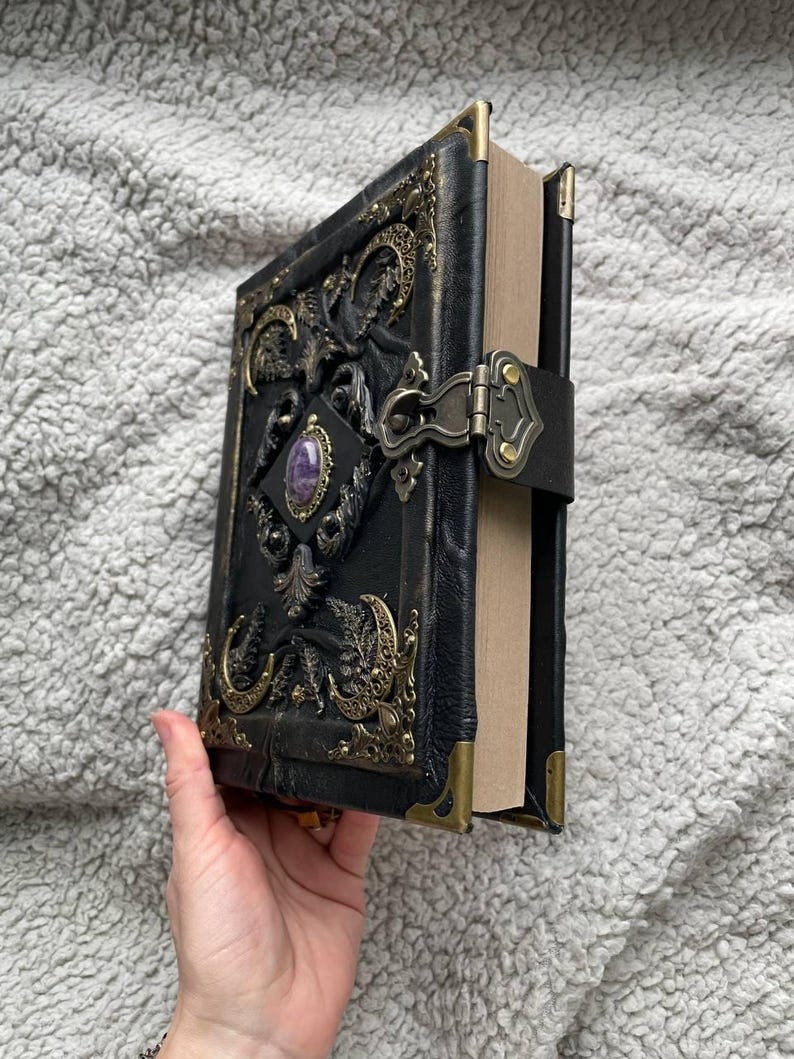 May include: A black leather-bound journal with ornate gold-colored metal accents and a purple gemstone. The cover features embossed designs and a clasp closure. The journal's corners are reinforced with gold-colored metal.