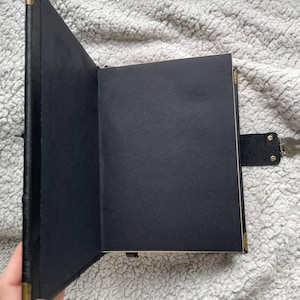 May include: Open black journal with blank black pages. The journal has a black cover with gold-colored corner protectors and a lock with a key. The journal is open, revealing the blank pages.