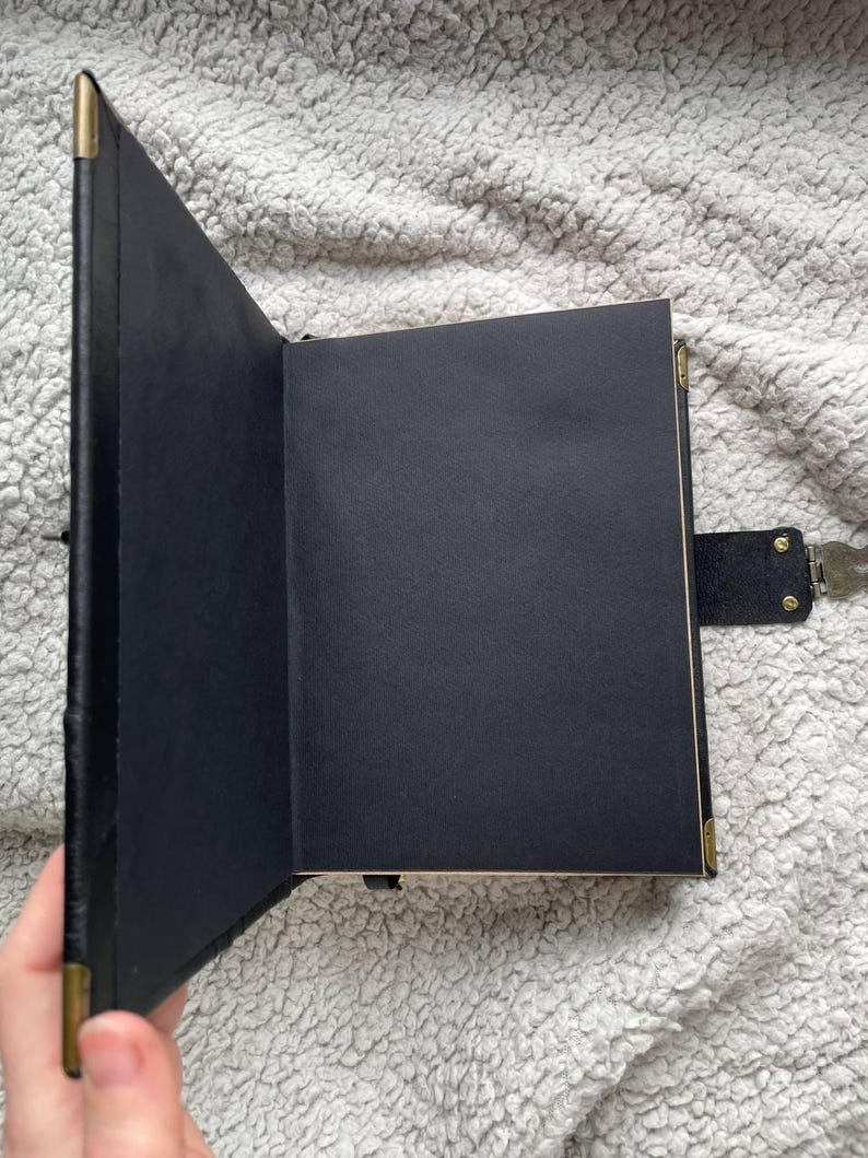 May include: Open black journal with blank black pages. The journal has a black cover with gold-colored corner protectors and a lock with a key. The journal is open, revealing the blank pages.
