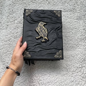 Pu&ograve; includere: Diario in pelle nera con un design a corvo in rilievo e dettagli decorativi agli angoli. Il diario ha una copertina strutturata e un emblema di corvo color oro. Sono visibili segnalibri a nastro neri.