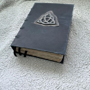 May include: A dark gray leather-bound book with a silver-colored Celtic knot emblem. The book has aged, yellowed pages and black ribbon bookmarks. The cover is textured, and the book appears to be handmade.