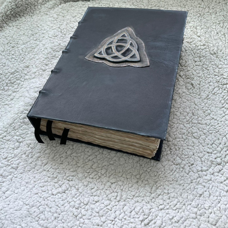 May include: A dark gray leather-bound book with a silver-colored Celtic knot emblem. The book has aged, yellowed pages and black ribbon bookmarks. The cover is textured, and the book appears to be handmade.