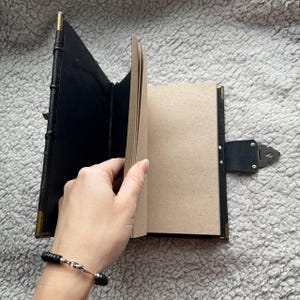 May include: Black leather journal with brass corner accents and a decorative clasp. The journal's pages are a light brown color, and the cover is open, revealing the interior pages. The journal is held open by a hand.
