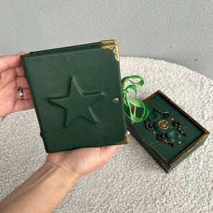 May include: A dark green leather-bound journal featuring an embossed star on the front cover, gold-toned metal corner protectors, and a green ribbon tie closure. Beside it sits a matching rectangular green box with ornate gold-toned metal accents.