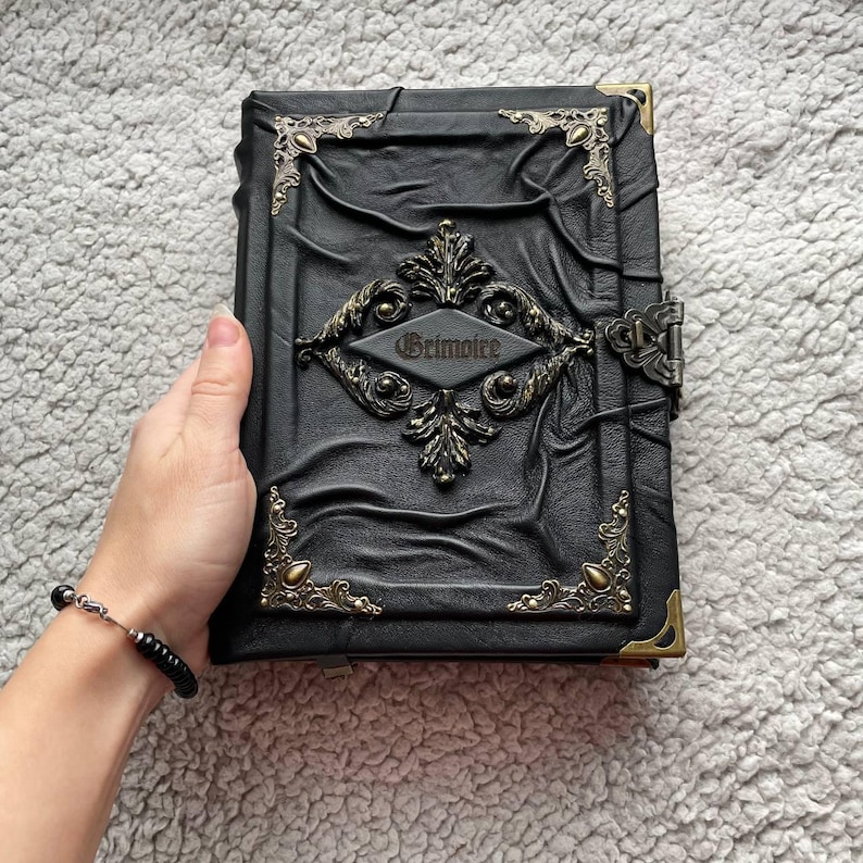 May include: A black leather-bound journal with ornate metal corner accents and a central decorative emblem. The word "Grimoire" is embossed on the front. The journal has a clasp closure.