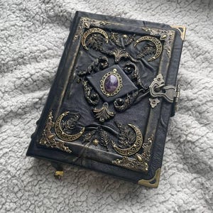 May include: Black leather journal with ornate brass accents. The cover features crescent moon designs, floral motifs, and a central amethyst stone. The journal has a clasp closure and brass corner protectors.