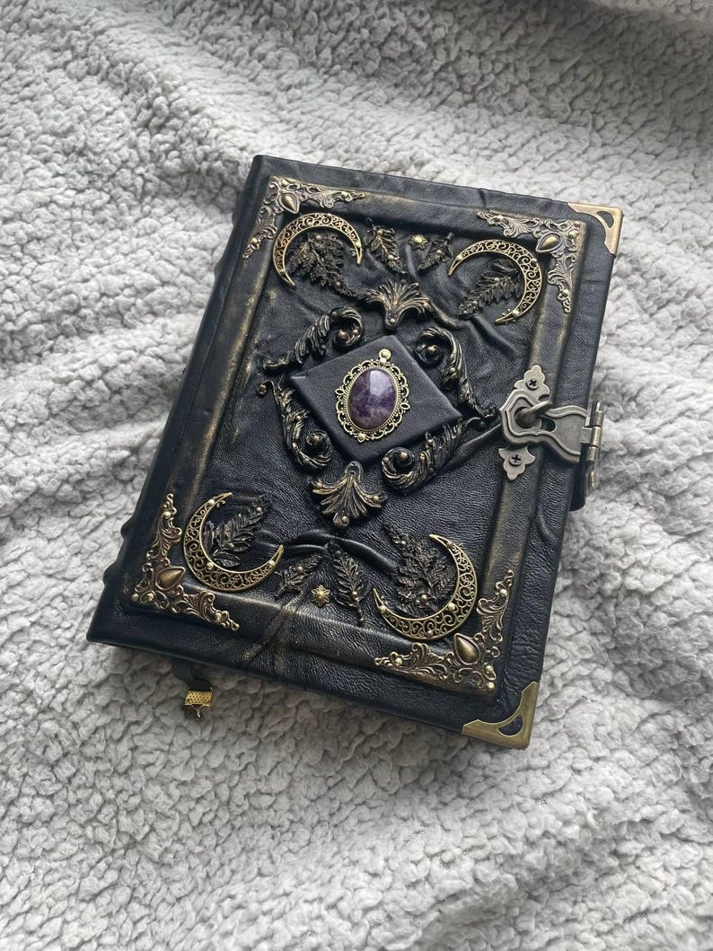 May include: Black leather journal with ornate brass accents. The cover features crescent moon designs, floral motifs, and a central amethyst stone. The journal has a clasp closure and brass corner protectors.