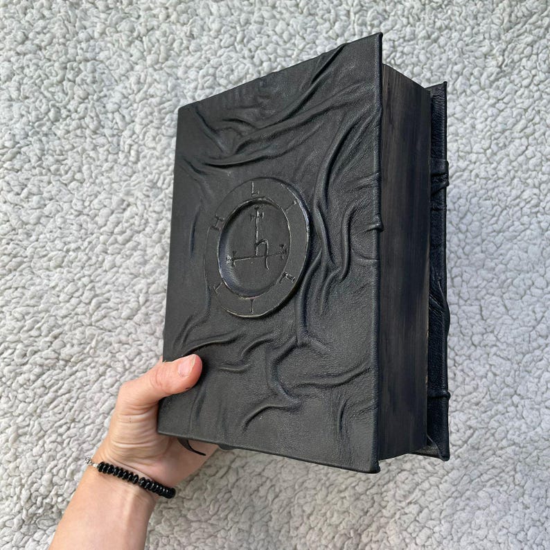 May include: A black, textured book with a circular emblem featuring a clock design. The book's cover has a rippled appearance. The book is held in a hand, and a black beaded bracelet is visible on the wrist.