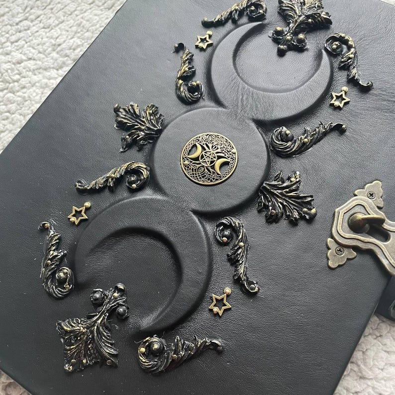 May include: Black leather journal with embossed crescent moons, stars, and ornate bronze-colored embellishments. Features a decorative circular emblem and a clasp closure. A gothic-themed notebook.