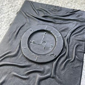 May include: A dark gray, leather-bound book with a circular emblem on the cover. The emblem features a cross and other symbols within a circle. The leather has a textured, wrinkled appearance.