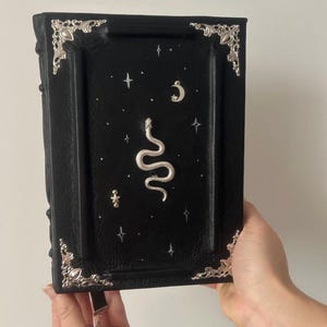 Op de afbeelding: Een zwart lederen gebonden dagboek met zilveren accenten en een decoratief ontwerp van een slang, sterren en een halve maan.