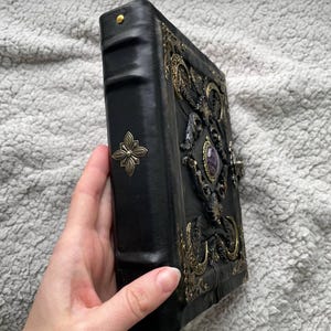 May include: Black leather-bound journal with ornate brass embellishments and a central purple gemstone. The cover features intricate relief designs and a decorative clasp. A brass flower adorns the spine.