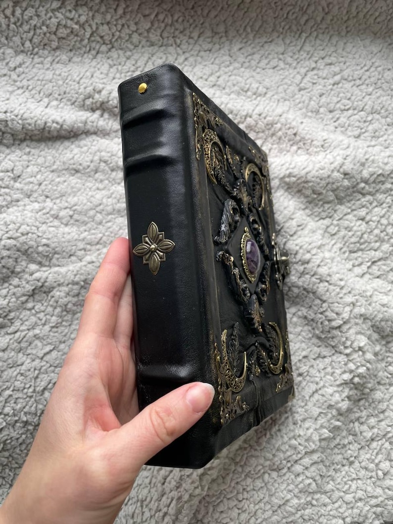 May include: Black leather-bound journal with ornate brass embellishments and a central purple gemstone. The cover features intricate relief designs and a decorative clasp. A brass flower adorns the spine.