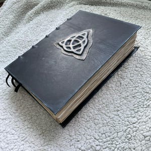 May include: A black leather-bound book with a silver Celtic knot emblem on the cover. The book has aged, off-white pages and a black ribbon bookmark. The cover is stitched along the spine.