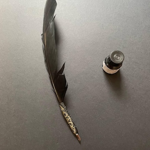 May include: A black quill pen with a gold filigree design on the handle, next to a small ink bottle. The feather is dark and angled, with a decorative metal band. The ink bottle is black with a white label.