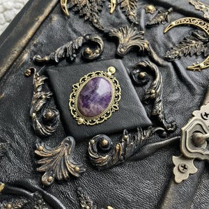 May include: A black leather-bound journal with ornate brass embellishments. The cover features a central square with a purple gemstone in a gold filigree setting. Decorative brass leaves, moons, and swirls adorn the cover.