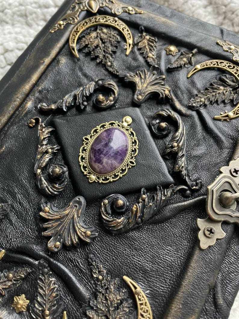 May include: A black leather-bound journal with ornate brass embellishments. The cover features a central square with a purple gemstone in a gold filigree setting. Decorative brass leaves, moons, and swirls adorn the cover.