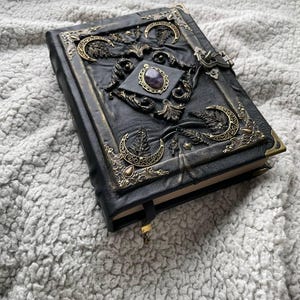 May include: Black leather journal with ornate brass accents. The cover features a central amethyst stone, crescent moon designs, and decorative corner pieces. A gold-colored ribbon bookmark is visible.