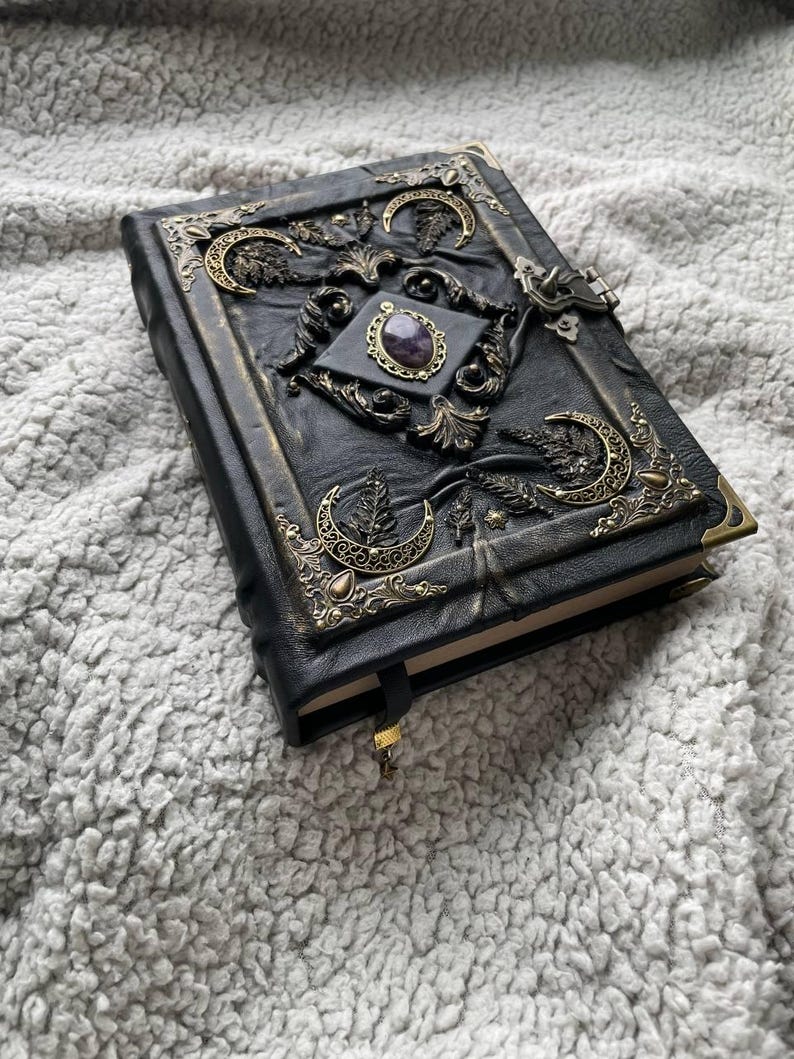 May include: Black leather journal with ornate brass accents. The cover features a central amethyst stone, crescent moon designs, and decorative corner pieces. A gold-colored ribbon bookmark is visible.