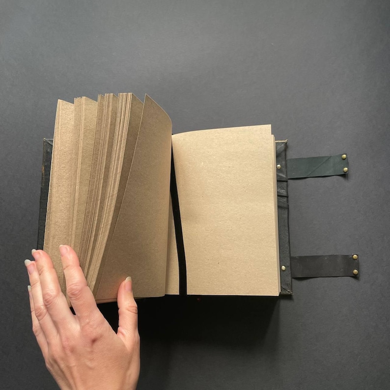 K&ouml;nnte beinhalten: Offenes Notizbuch mit Kraftpapierseiten und dunklem Ledereinband. Das Notizbuch wird mit zwei Lederriemen und Messingnieten verschlossen. Eine Hand bl&auml;ttert die Seiten um.